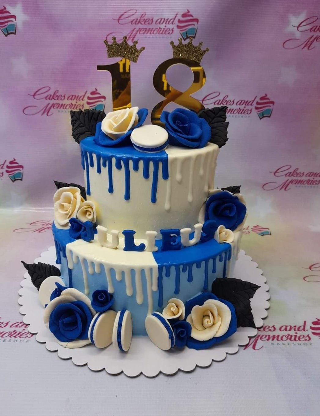 Two-tier blue and white cake with blue roses, white macarons, and gold number 18 toppers with crowns.