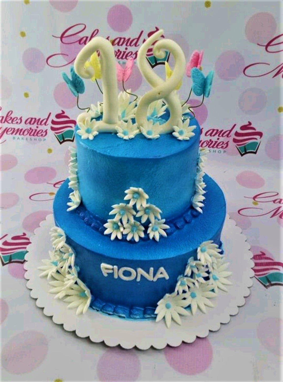 Two-tier blue birthday cake with white fondant daisies and large white number 18 topper with butterflies.