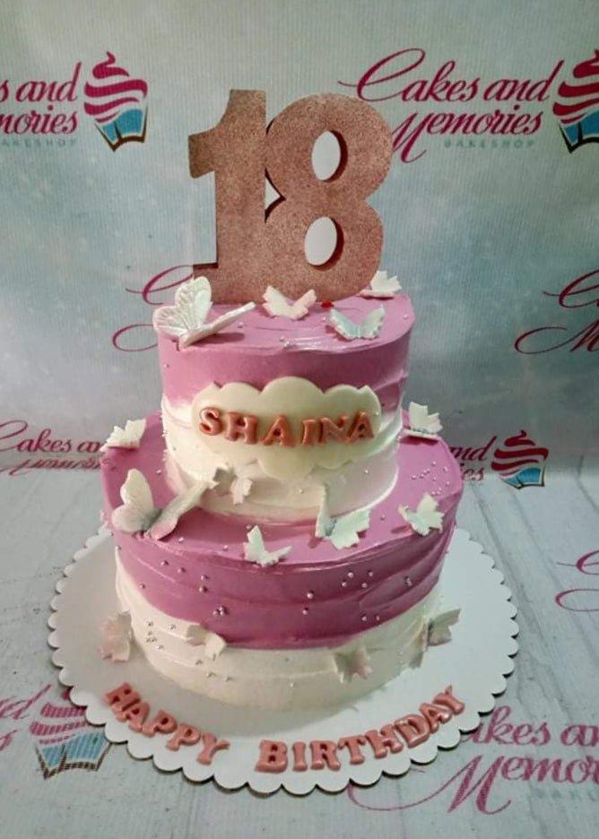 Two-tier pink and white butterfly birthday cake with a large rose gold glitter number 18 topper and name Shaina on a cloud.