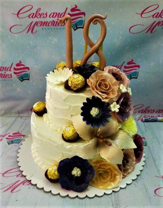 Two-tier white soft-icing cake decorated with large gumpaste flowers in purple, tan, and yellow, topped with a large number 18 and Ferrero chocolates.
