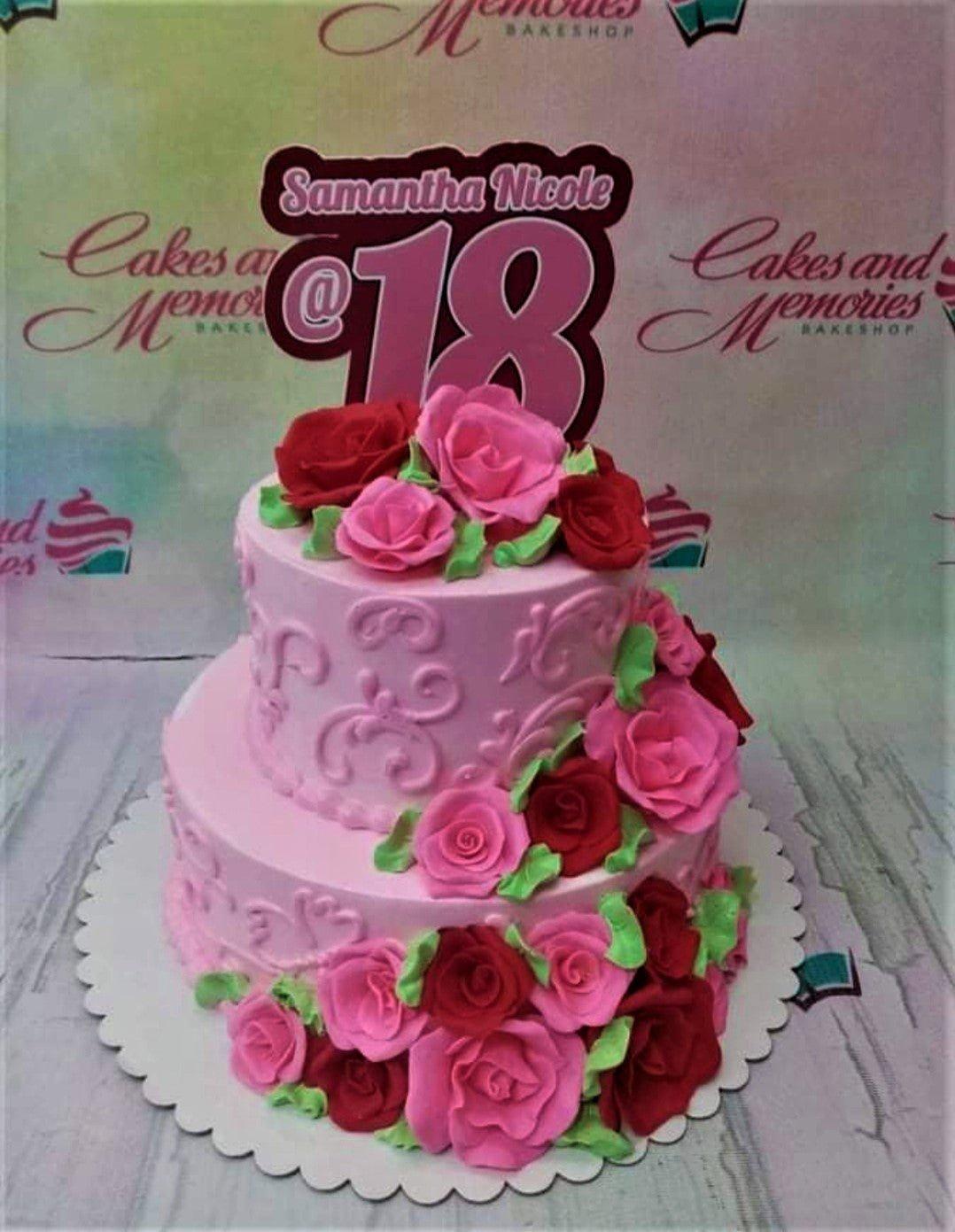 Two-tier pink cake decorated with red and pink gumpaste roses and a large 'Samantha Nicole @ 18' printout topper.