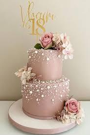 A pink 2-tier cake featuring gold '18' and name toppers, pink roses, white hydrangea clusters, and white pearl sprinkles.