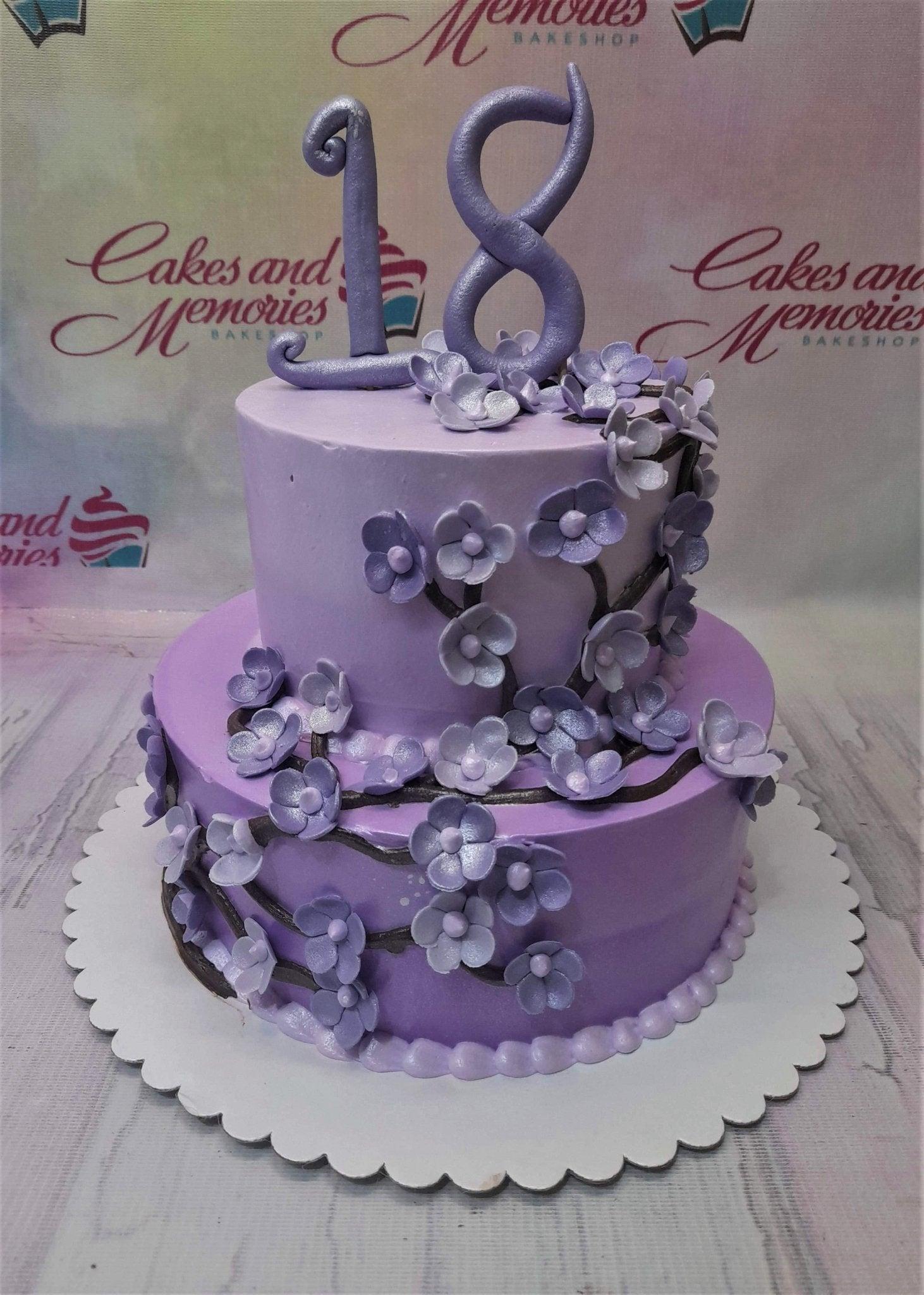 Two-tier lavender purple cake decorated with edible purple flowers on brown branches and a large purple number 18 topper.