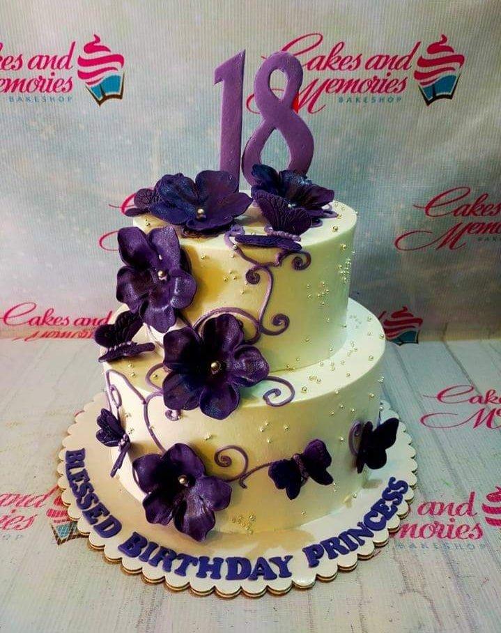 Two-tier white cake with purple fondant flowers, butterflies, and a large purple number 18 topper.