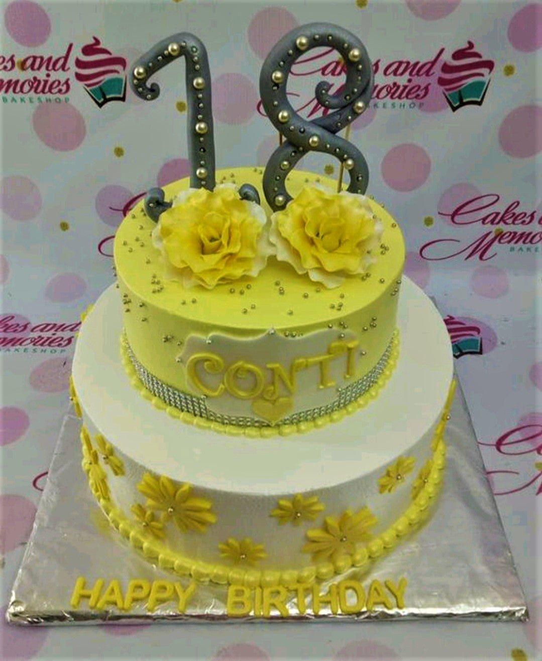 Two-tier yellow and white birthday cake with large grey number 18 toppers, yellow roses, and silver dragees.