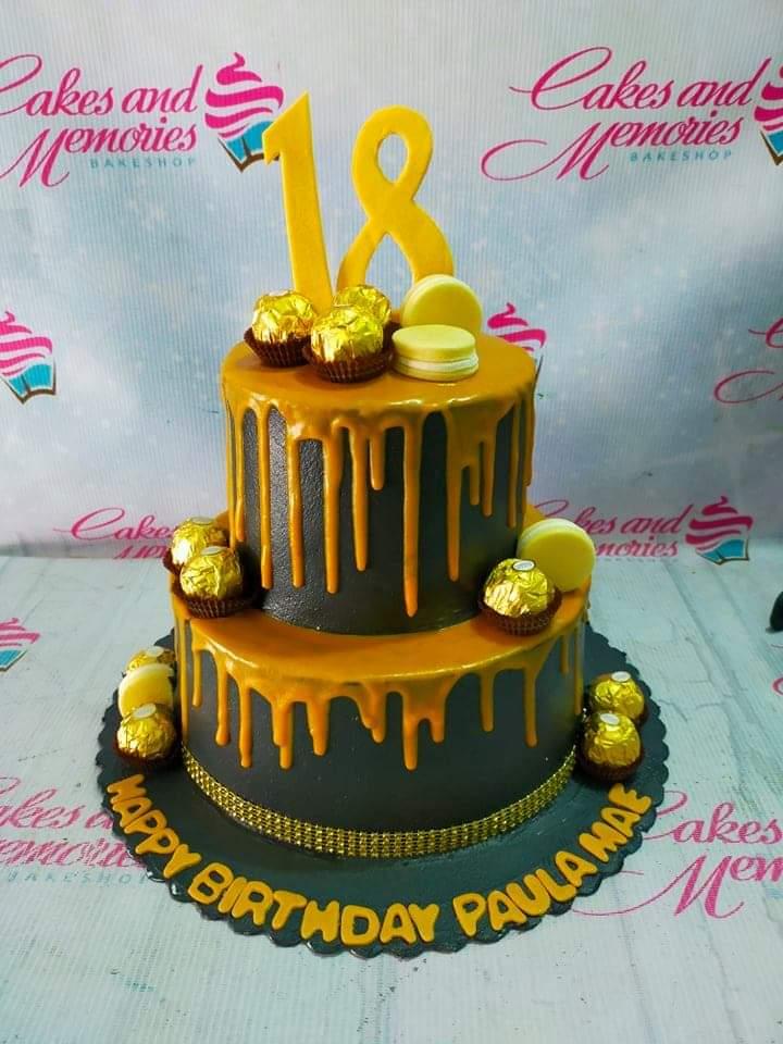 Two-tier black and gold drip cake with Ferrero Rocher chocolates, macarons, and a large yellow number 18 topper.