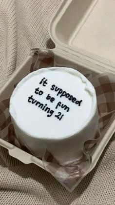 White bento cake with black piped icing text in a brown checkered liner inside a takeout box.