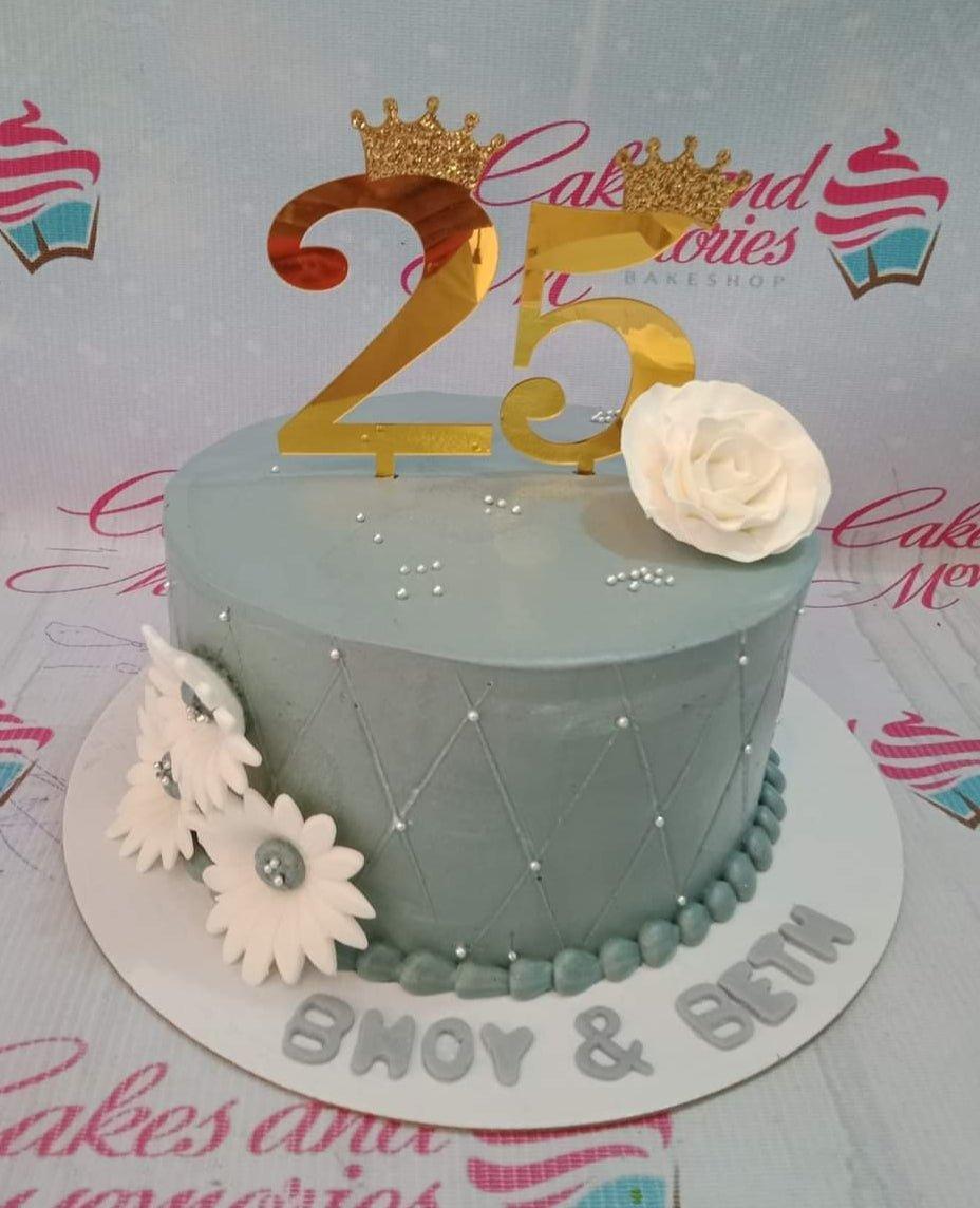 A single-tier gray cake with a quilted pattern, white fondant flowers, and gold number 25 toppers with crowns.