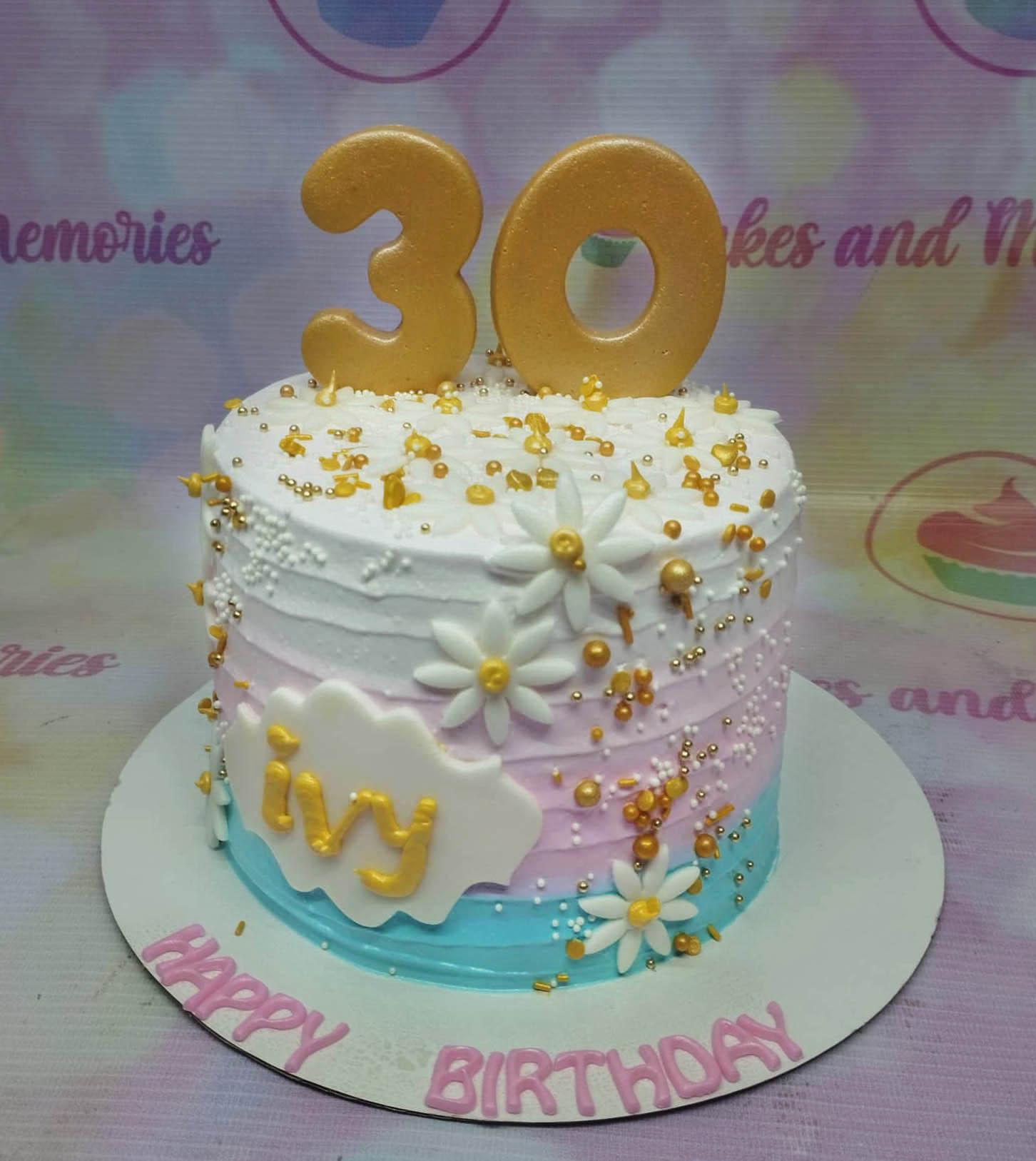 A single-tier birthday cake with white, pink, and blue gradient icing, gold number 30 toppers, white daisies, and gold sprinkles.