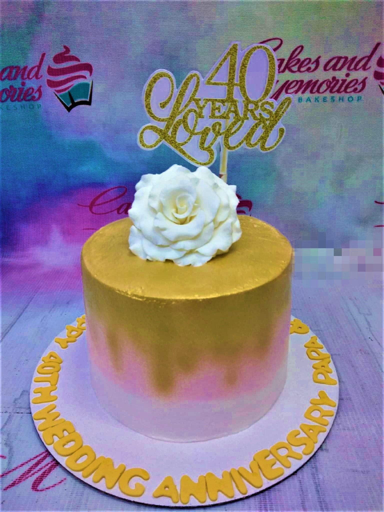 A single-tier 40th wedding anniversary cake with a gold, pink, and white gradient icing, topped with a large white edible rose and a gold glitter topper.