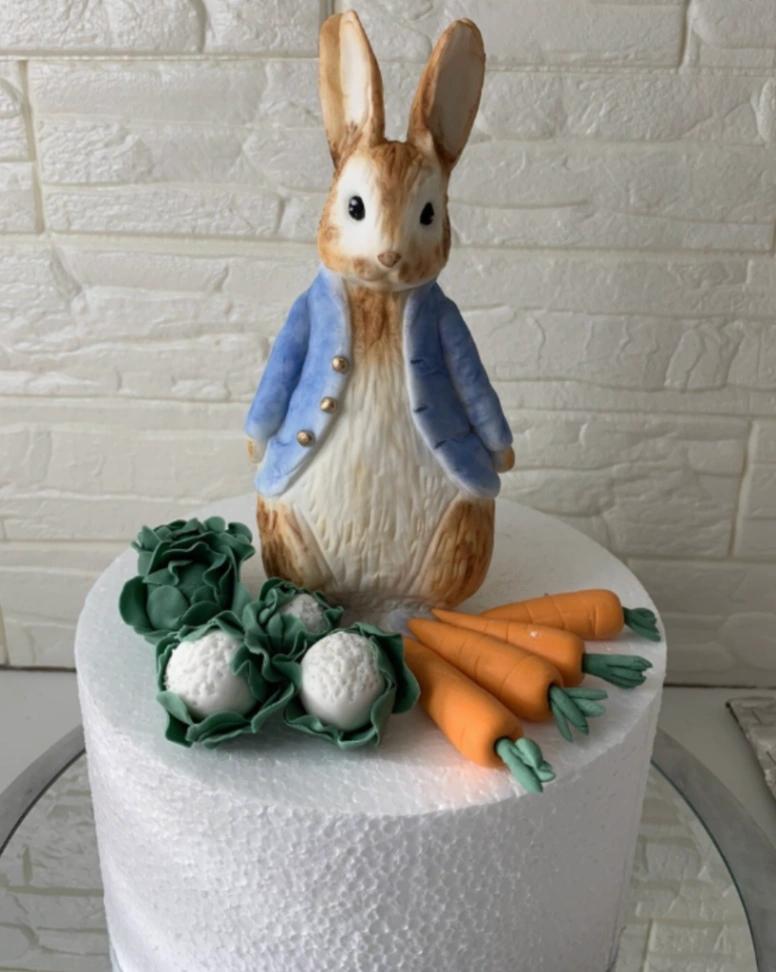 Peter Rabbit themed fondant cake with a large 3D edible rabbit figure, fondant carrots, and cabbages on a white base.