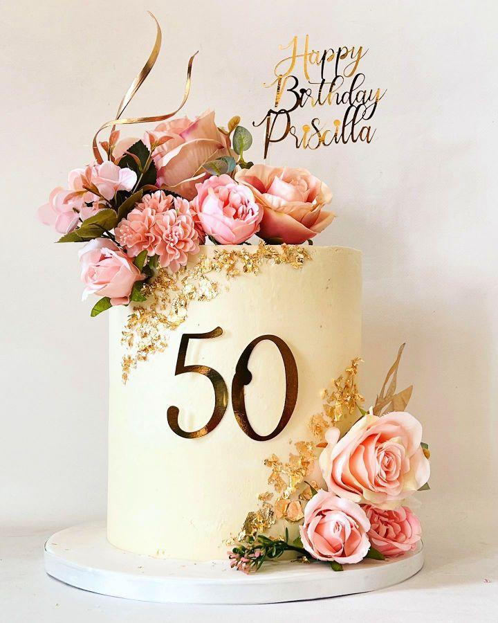 Elegant cream-colored tall cake for a 50th birthday featuring pink and peach artificial roses, gold leaf accents, and a gold acrylic topper.