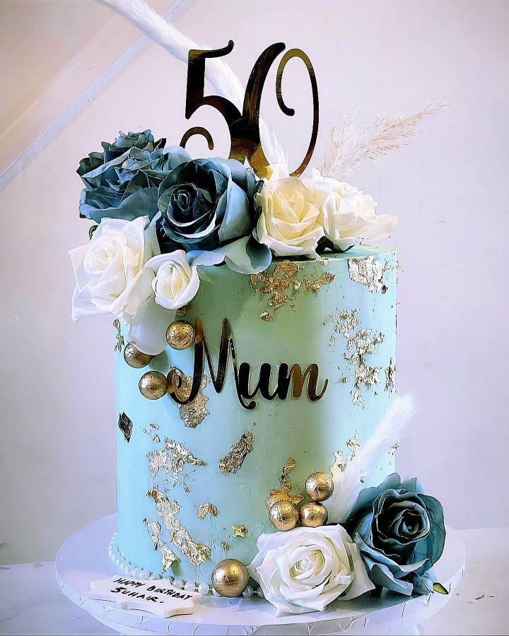 Tall mint green cake with gold leaf accents, blue and white artificial roses, gold spheres, and a gold 50 topper.