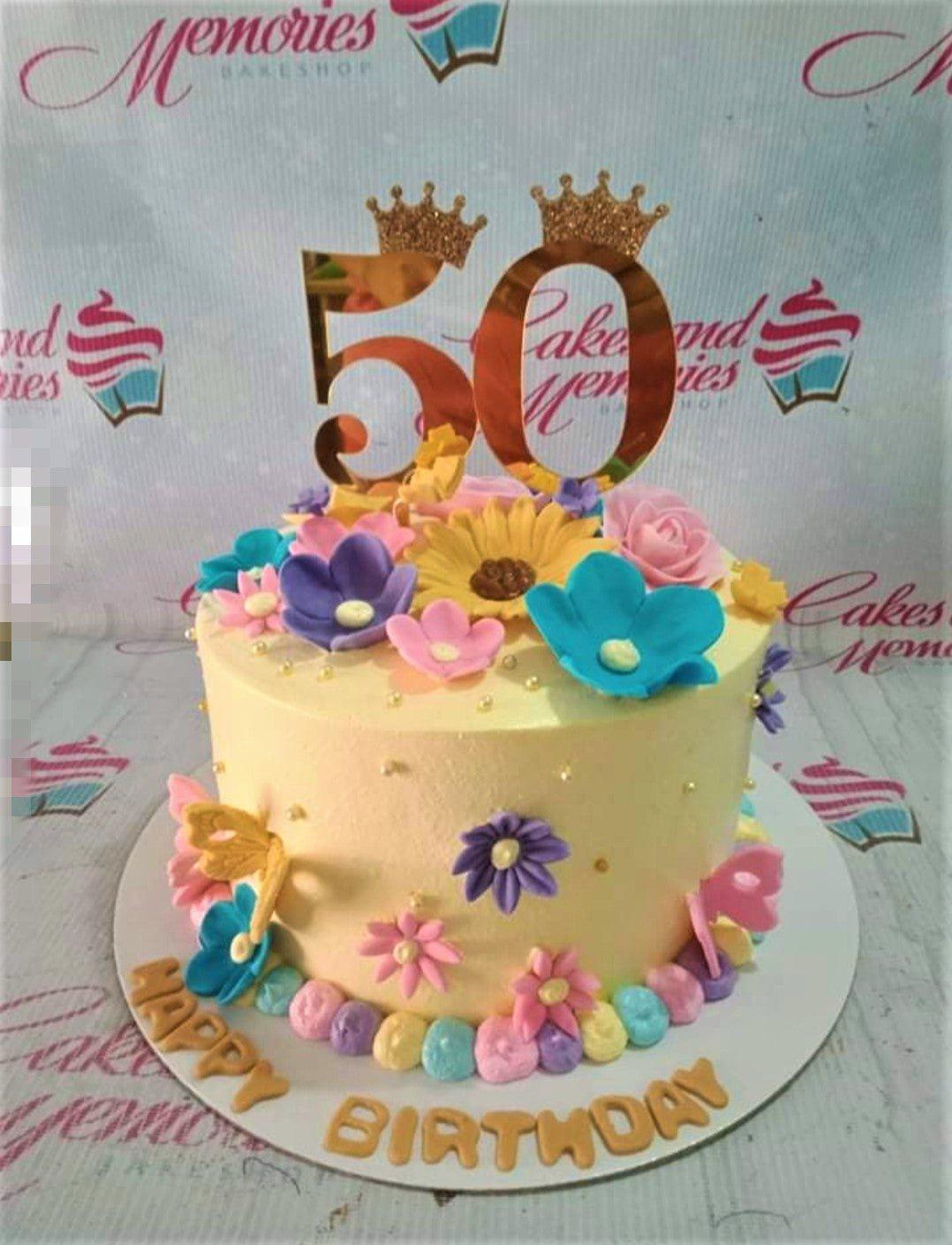 Yellow 50th birthday cake decorated with colorful fondant flowers, butterflies, and a gold number 50 topper with crowns.