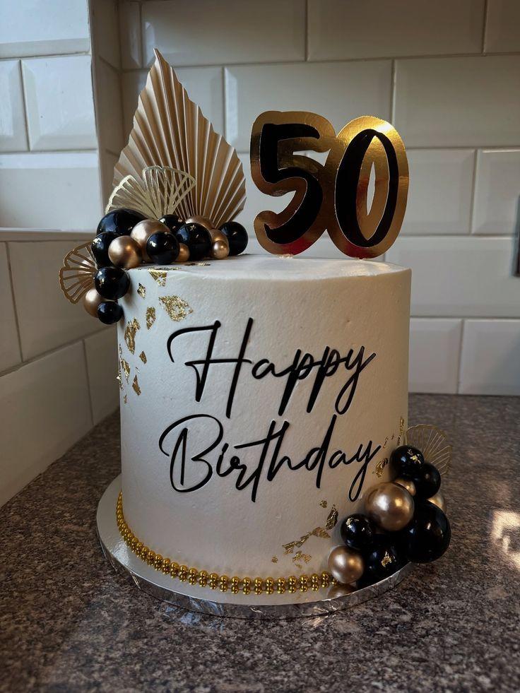 White 1-tier 50th birthday cake with gold and black balloon toppers, gold palm leaves, and black 'Happy Birthday' text.