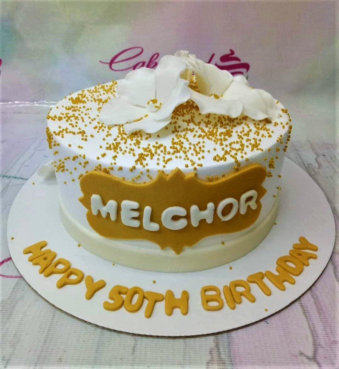 White 1-tier birthday cake with gold dragees, white gumpaste flowers, and a gold name plaque for a 50th birthday celebration.