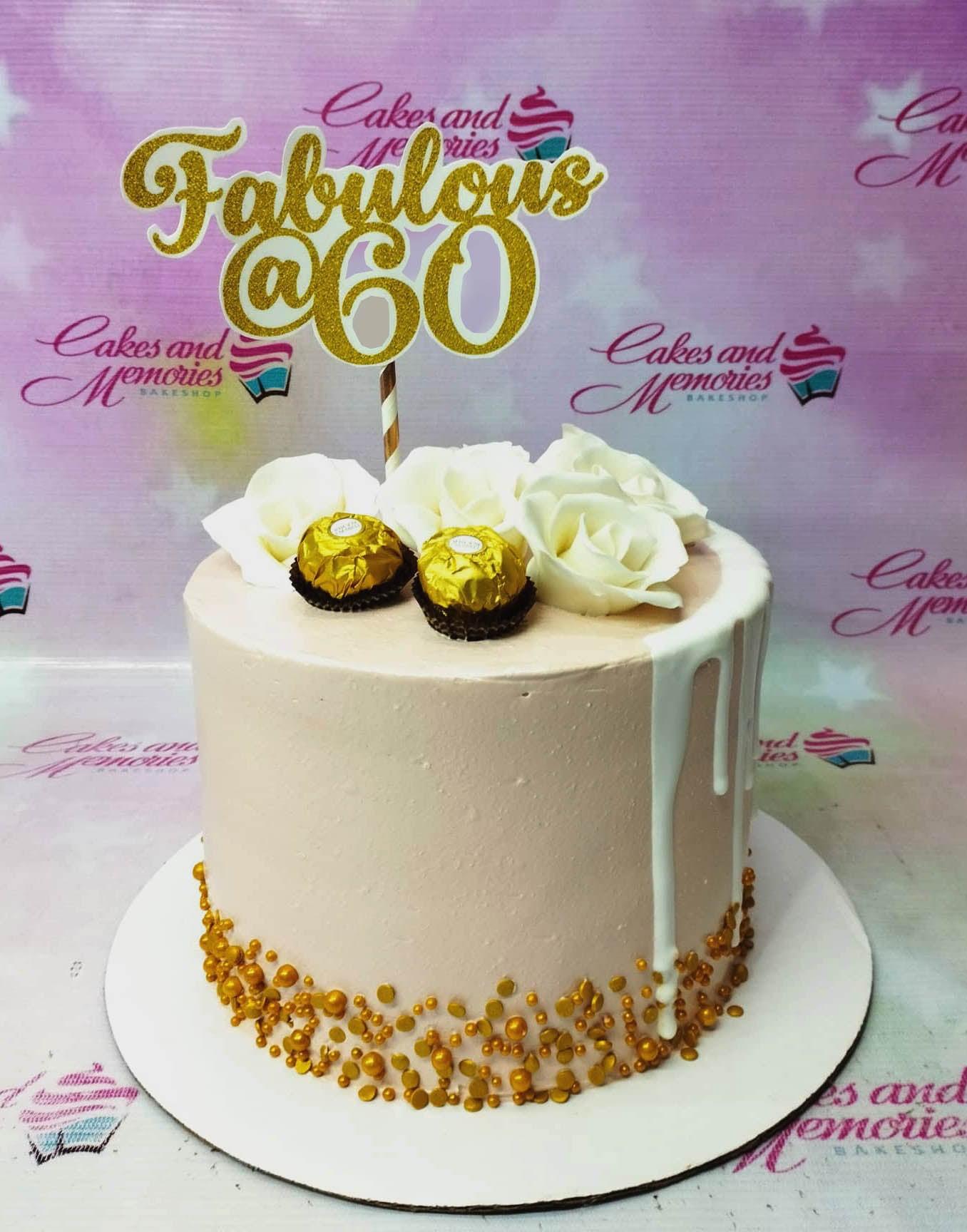 Elegant light pink 60th birthday cake with white roses, Ferrero Rocher chocolates, gold dragees, and a gold glitter Fabulous @ 60 topper.