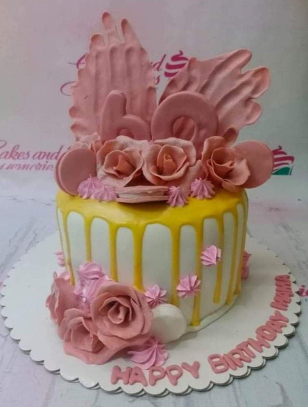 White 1-tier cake with yellow drip, pink icing rosettes, and large pink gumpaste roses and wings for a 60th birthday.