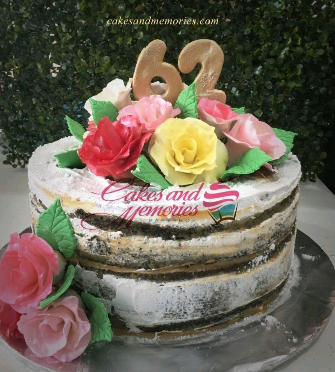 Naked chocolate cake with white icing, topped with large gold number 62 and a bouquet of pink, red, and yellow edible roses.