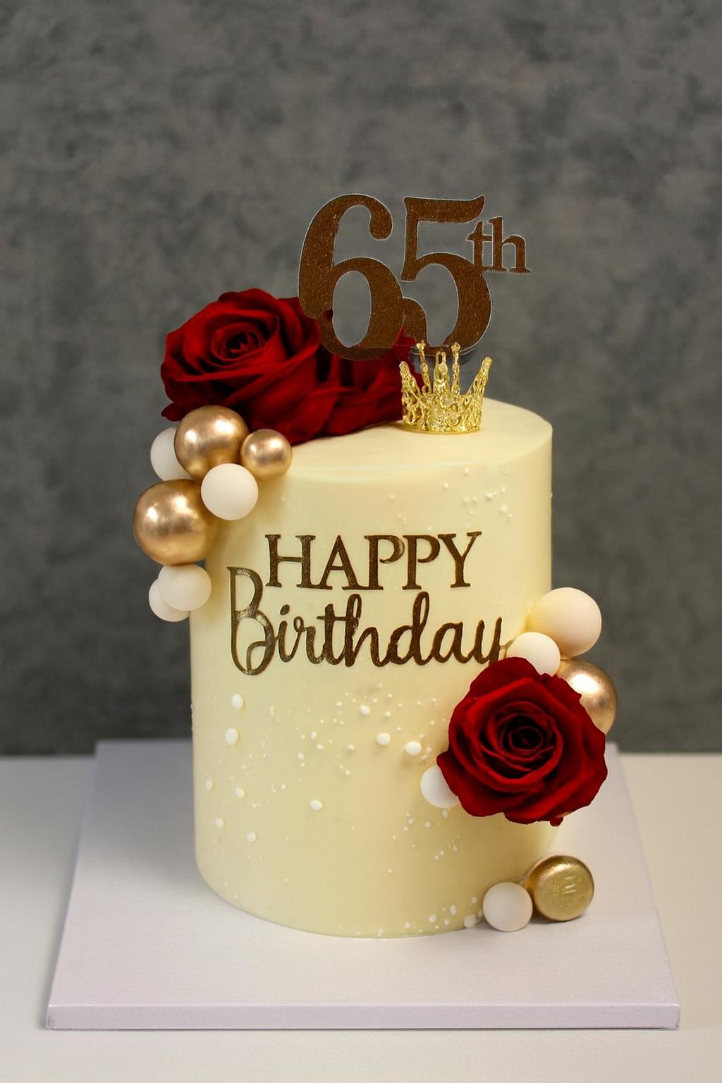 Tall ivory buttercream cake for a 65th birthday featuring red roses, gold and white spheres, a gold crown, and a 65th topper.