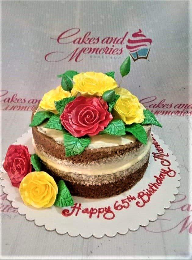 Naked chocolate cake with yellow and red gumpaste roses and green leaves, featuring a Happy 65th Birthday message on the base board.