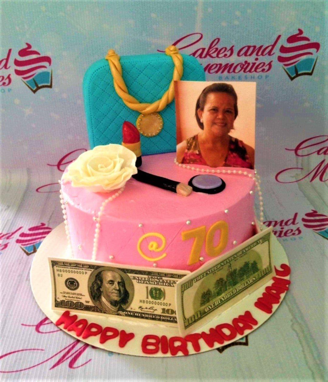 Pink 70th birthday cake with a blue fondant handbag, makeup toppers, a photo printout, and edible dollar bills.