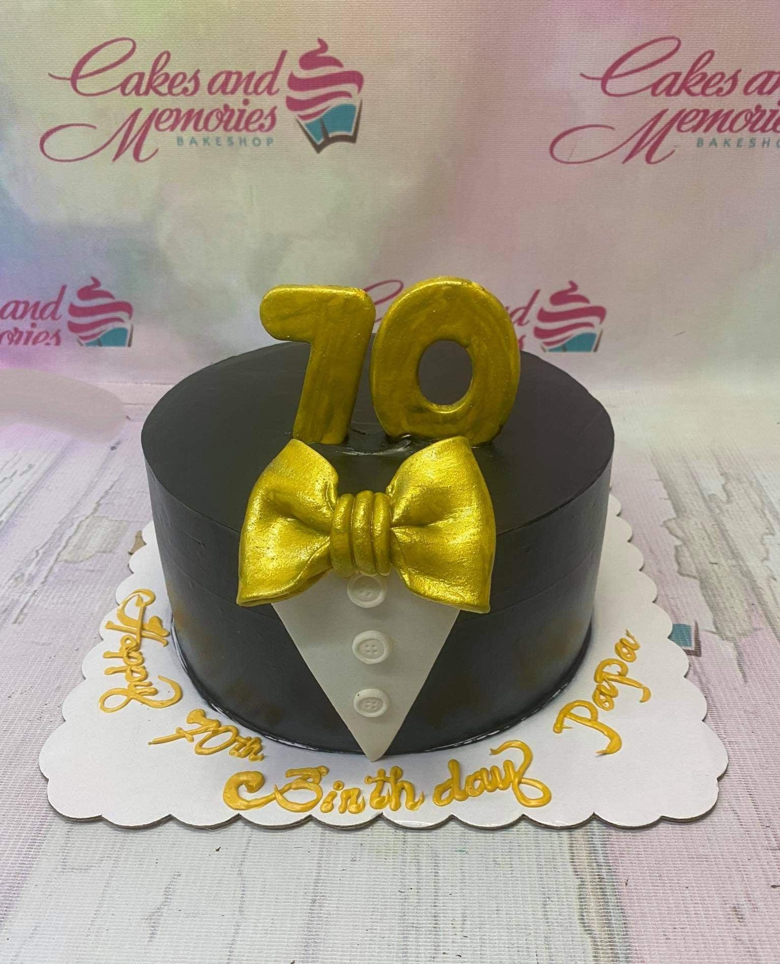 Black tuxedo themed 70th birthday cake with gold number 70 and bow tie toppers on a white board with gold icing message.