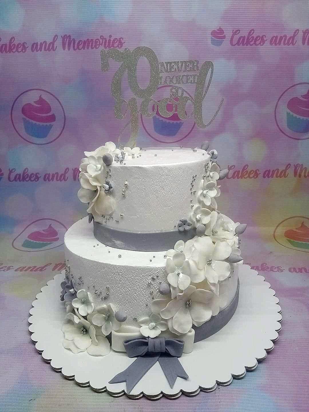Elegant white 2-tier 70th birthday cake with white gumpaste flowers, silver dragees, and a silver glitter topper.