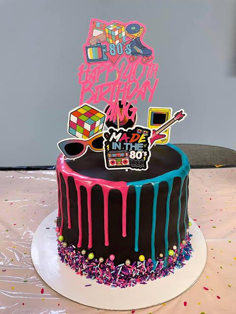 Black 80s themed birthday cake with neon pink and blue drips, colorful sprinkles, and retro printout toppers like a Rubik's cube and boombox.