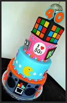 Vibrant 3-tier 80s themed birthday cake with Rubik's Cube top, Pac-Man, MTV logo, and a disco ball topper.