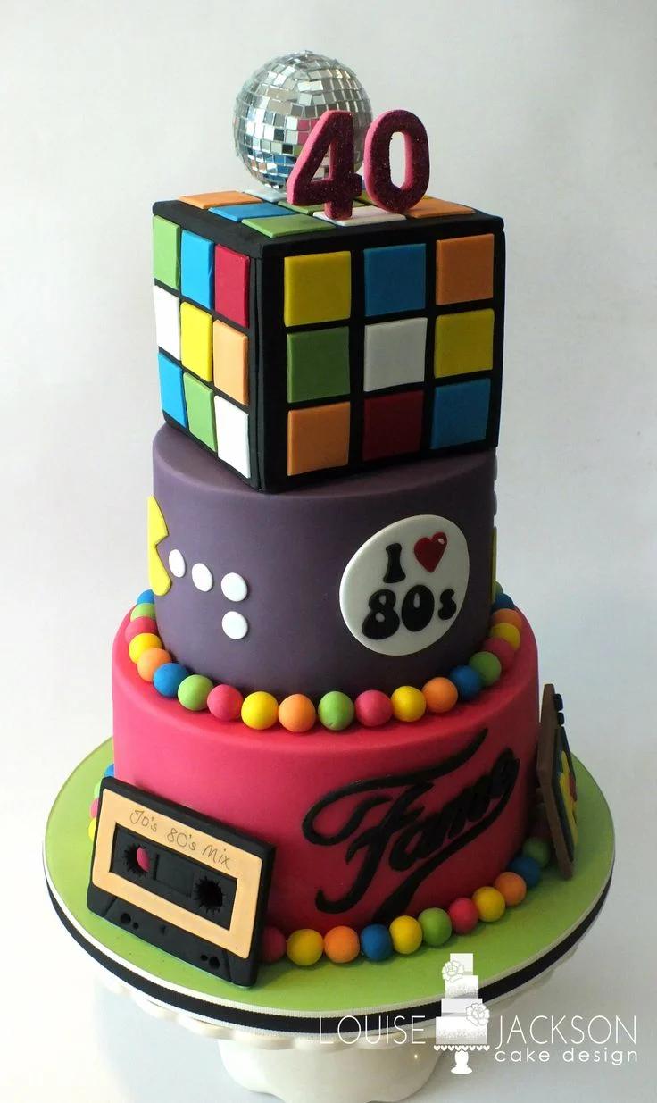 3-tier 80s themed fondant cake with Rubik's cube top, purple middle tier with Pac-Man, and pink bottom tier with cassette tape.