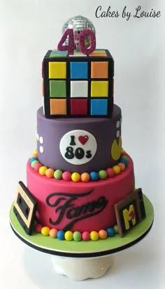 3-tier 80s themed cake with Rubik's cube top, purple and pink tiers, disco ball, and retro icons like Pac-Man and cassette tapes.