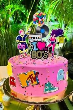 Vibrant pink 80s themed birthday cake with retro printouts of a Rubik's cube, cassette tape, MTV logo, and Pac-Man ghosts.