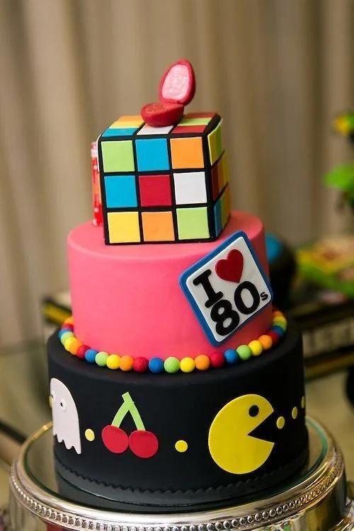 Two-tier 80s themed fondant cake with a Rubik's Cube topper, Pac-Man, and ghost decorations on black and pink tiers.