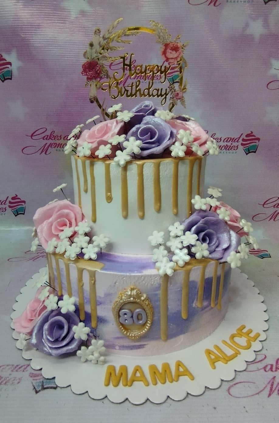 Two-tier purple and white cake with gold drips, pink and purple roses, and a gold Happy Birthday topper for an 80th birthday.