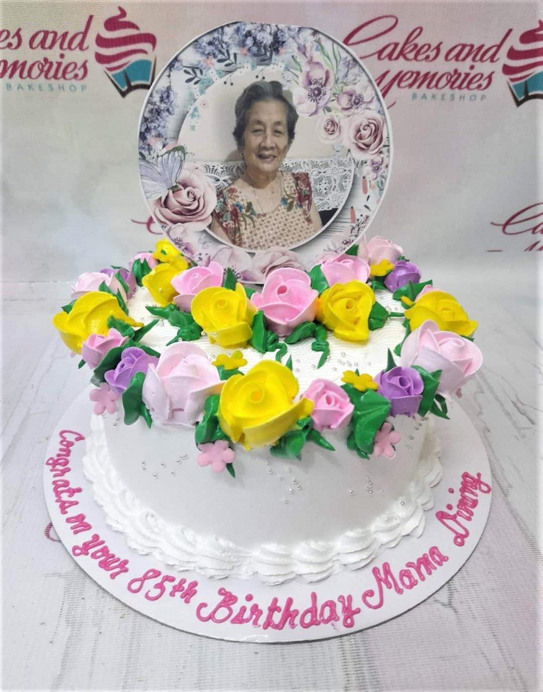 White 1-tier birthday cake for an 85th birthday featuring a photo printout topper and a wreath of pink, yellow, and purple icing roses.