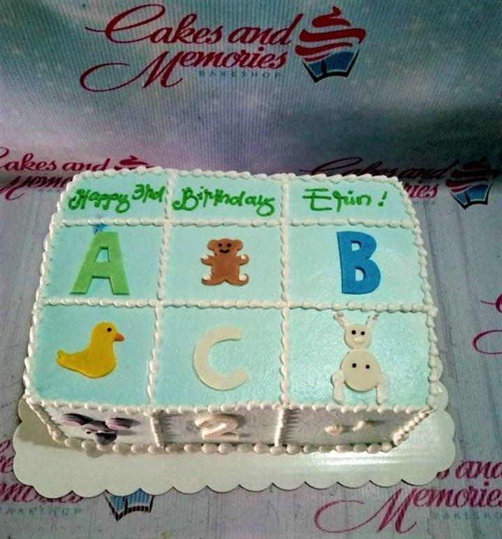 Light blue square birthday cake with white icing borders, featuring alphabet blocks, a teddy bear, a duck, and a bunny.
