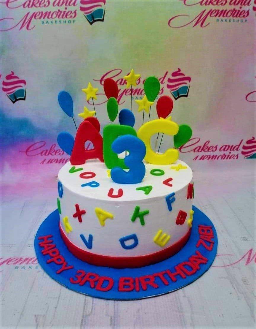 White 1-tier birthday cake with colorful fondant alphabet letters and 3D ABC balloon toppers on a blue base board.