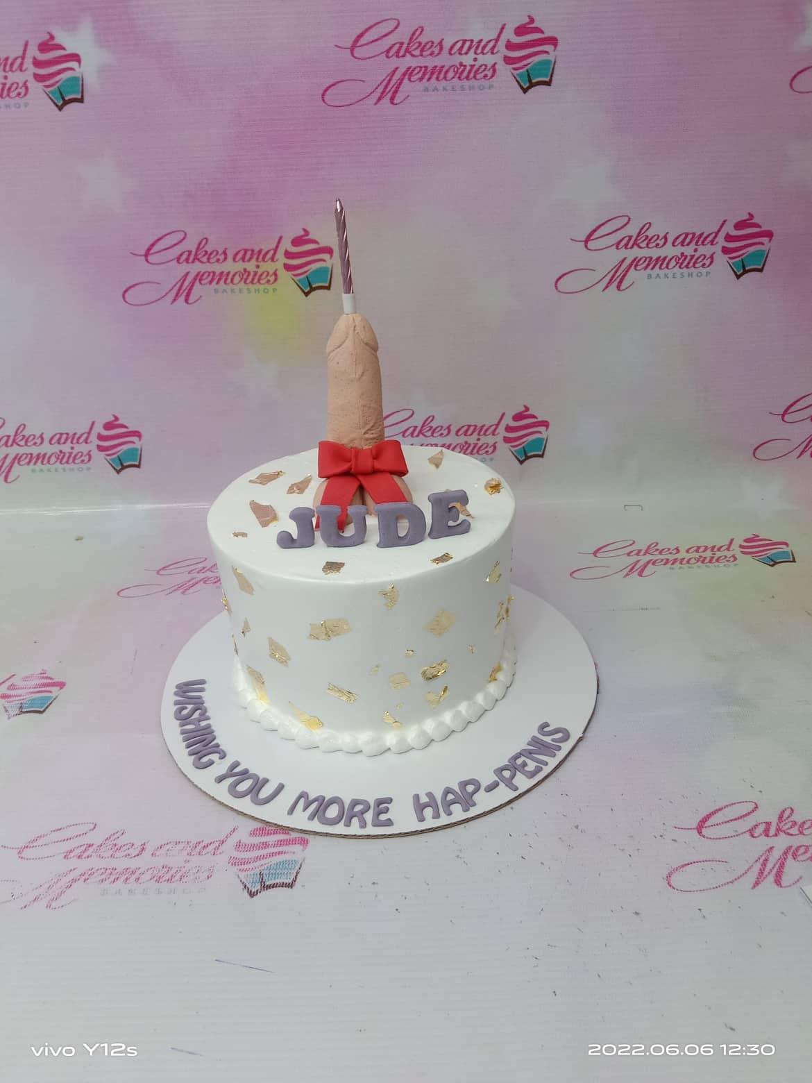 White minimalist cake with a humorous phallic topper, red bow, and gold leaf accents for a birthday celebration.