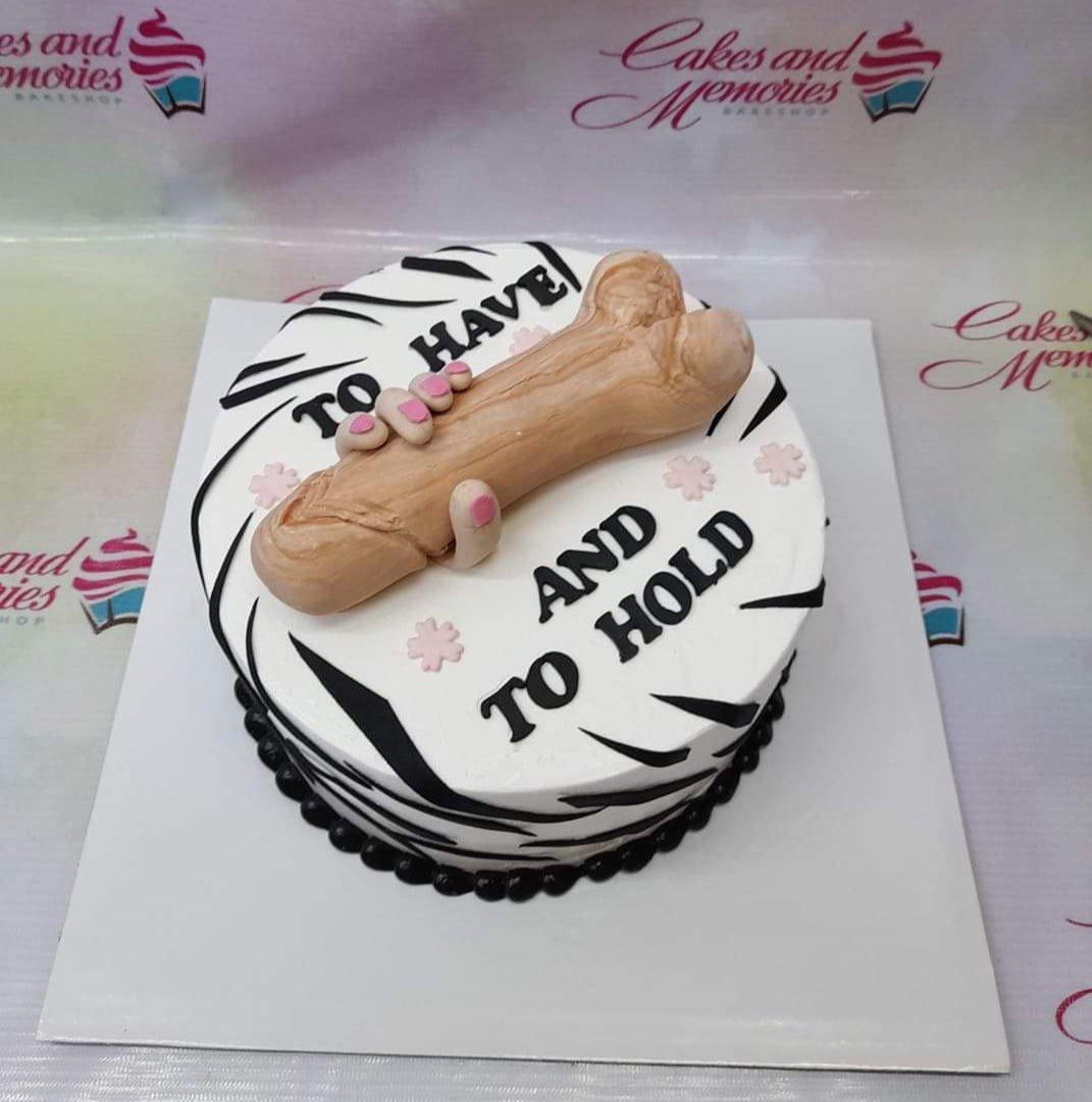 White round cake with black zebra stripes, pink flowers, and a large tan fondant phallic-shaped topper being held by a hand.