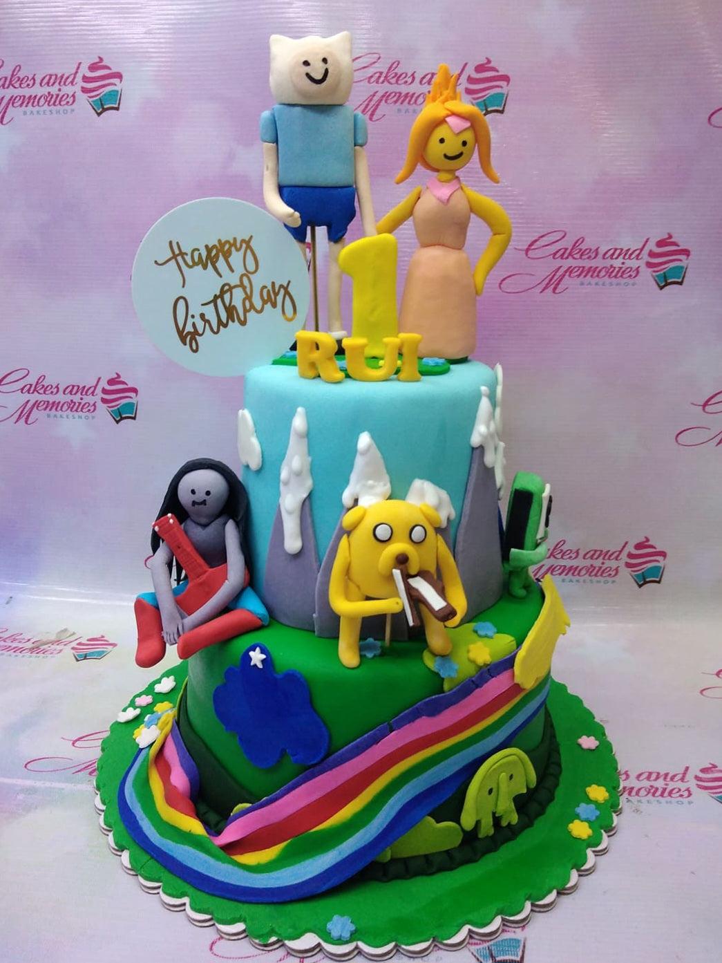 Adventure Time themed 2-tier fondant cake with Finn, Princess Bubblegum, Marceline, and Jake figures on a colorful landscape.