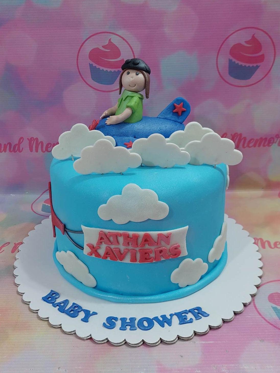 Blue fondant airplane themed baby shower cake with a 3D pilot figure, white clouds, and a red banner.