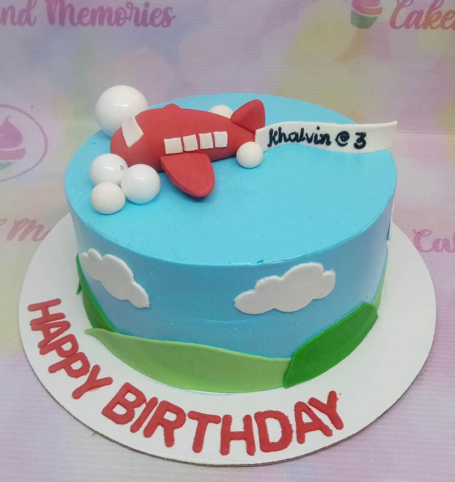 Light blue airplane themed birthday cake with a red fondant plane, white clouds, and green hills on a white base board.