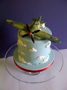 A light blue fondant cake featuring a 3D camouflage airplane topper flying through white fondant clouds.
