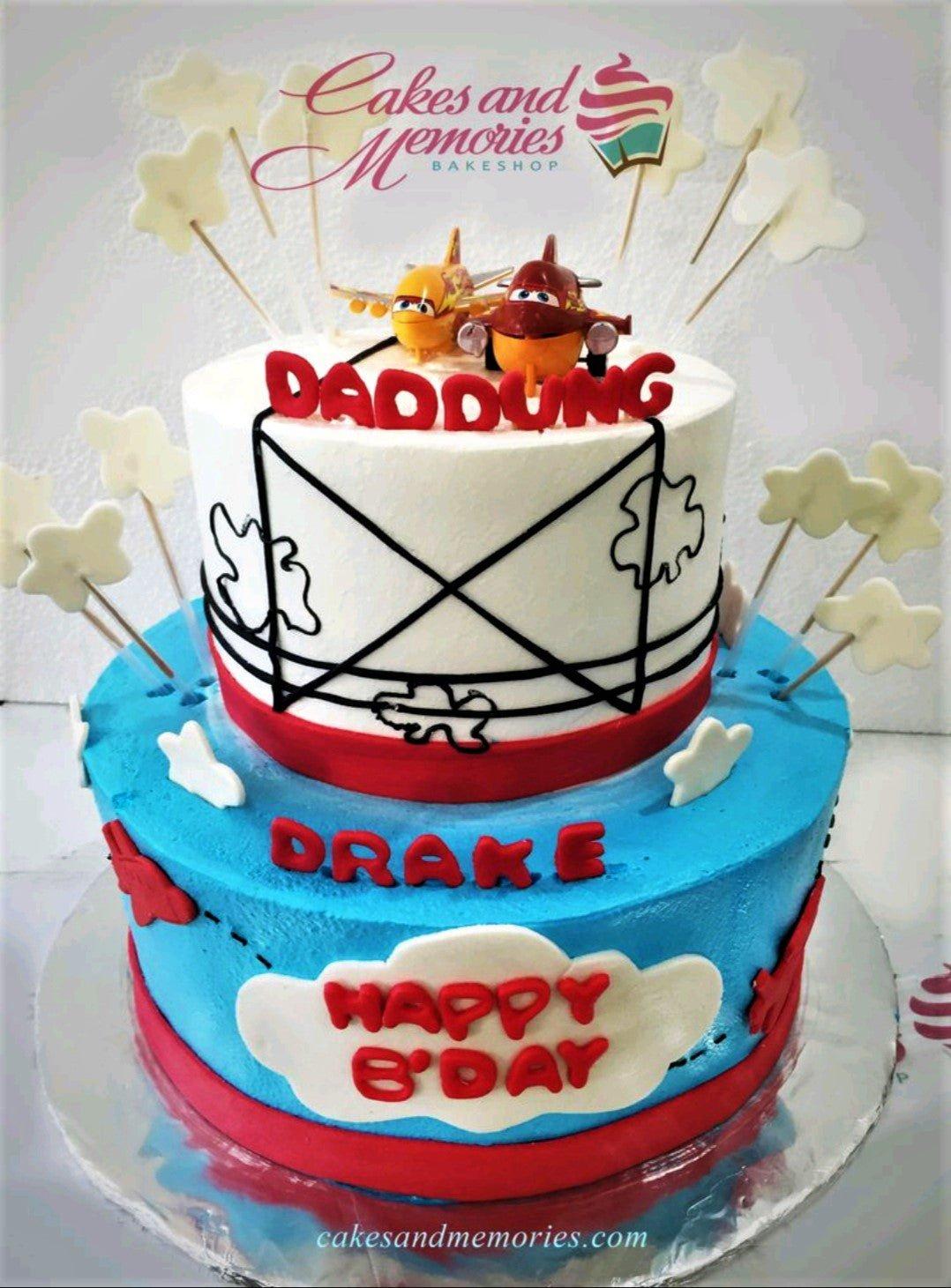 Two-tier airplane themed birthday cake in white and blue with red accents, featuring 3D plane figures and cloud decorations.