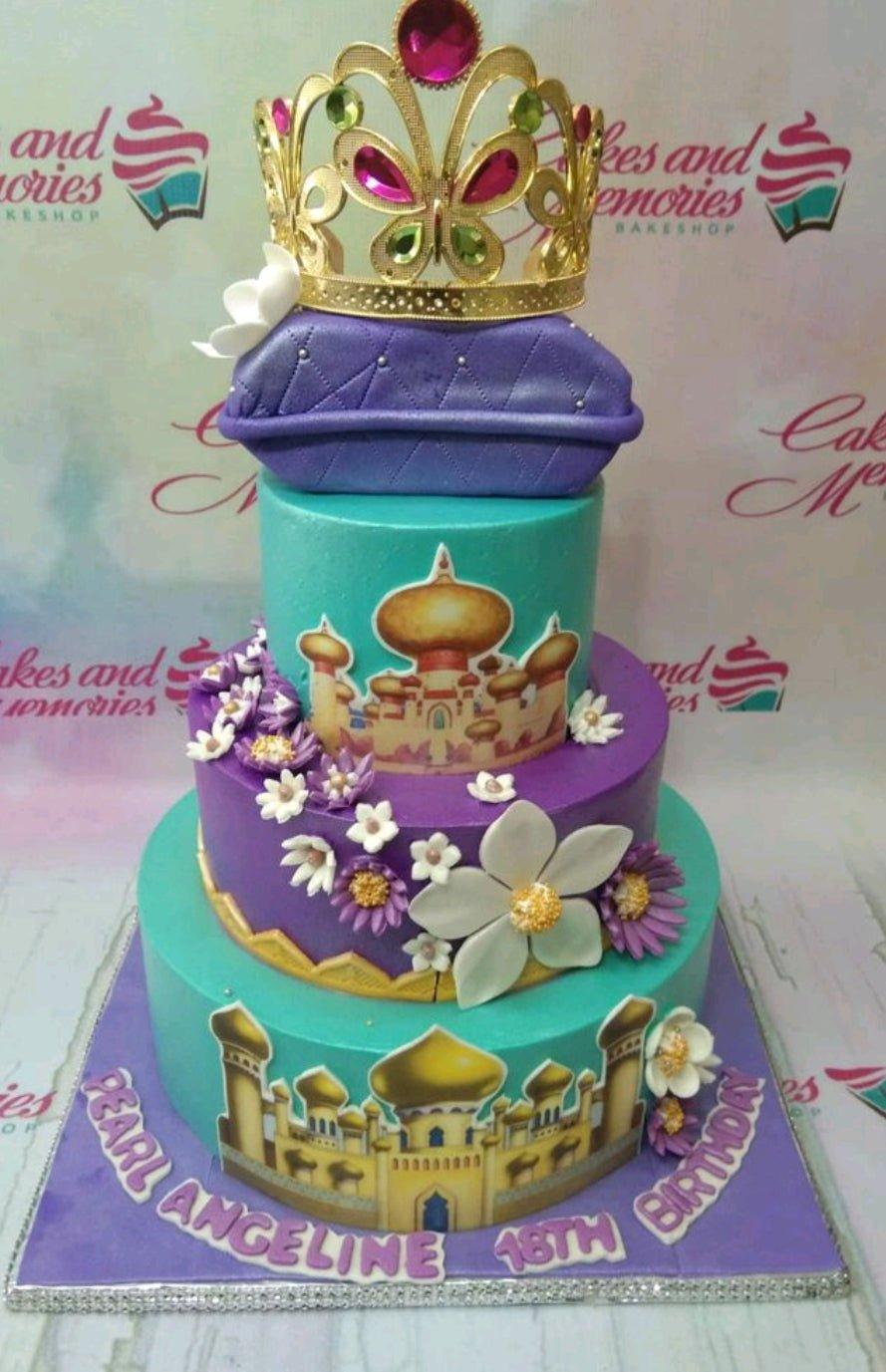 Three-tier teal and purple Aladdin themed cake with a gold crown on a purple pillow topper and palace printouts.