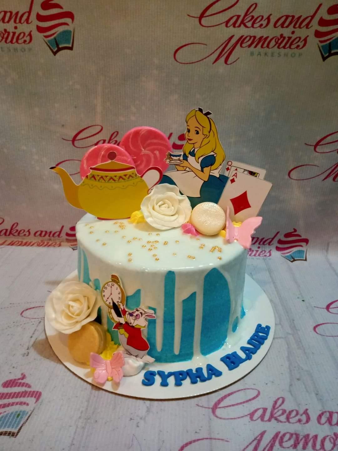 Alice in Wonderland themed cake with blue icing, white drip, Alice and White Rabbit printouts, and pink lollipops.