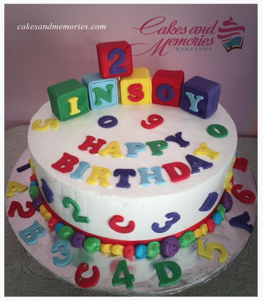White round birthday cake with colorful fondant alphabet blocks spelling INSOY and scattered numbers and letters on the top and sides.