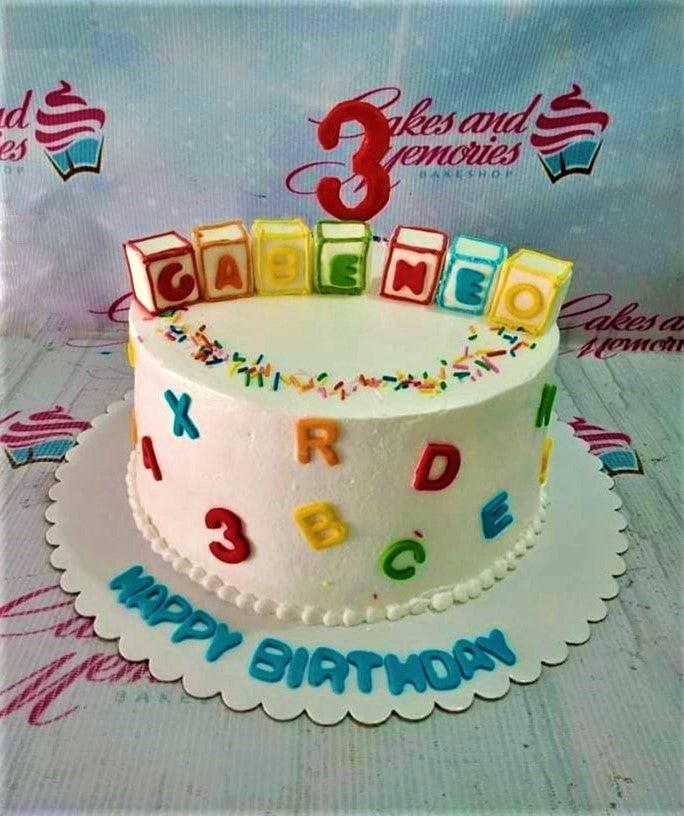White 1-tier birthday cake with colorful alphabet blocks, scattered letters, and rainbow sprinkles.