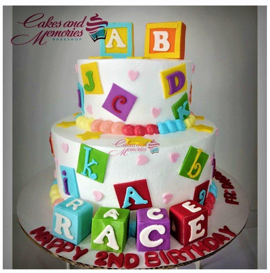 Two-tier white birthday cake decorated with colorful alphabet blocks and letter squares for a 2nd birthday celebration.
